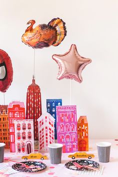 a table topped with paper plates and cups filled with cake next to balloon shaped buildings