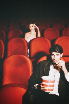 a man and woman sitting in the middle of an auditorium with red seats, one holding a drink