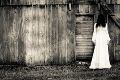 a woman standing in front of a wooden building with her back to the camera, wearing a long white dress