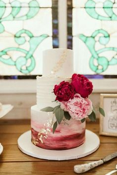 a three tiered cake with flowers on top sitting on a wooden table next to a stained glass window