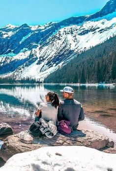 two people are sitting on the edge of a mountain lake