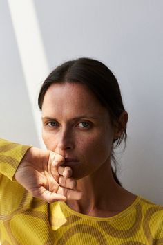 a woman in a yellow shirt holding her hand up to her face and looking at the camera