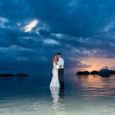 a bride and groom standing in the water at sunset on their wedding day with boats in the background