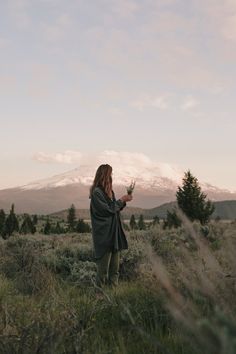 a woman is standing in the grass looking at her cell phone with a mountain in the background