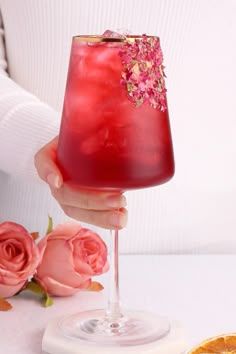 a person holding a wine glass with red liquid and pink flowers on the table next to it