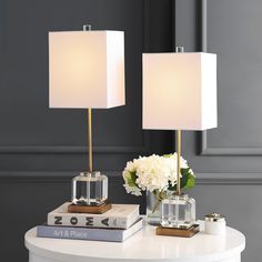 two lamps sitting on top of a white table next to books and vase with flowers