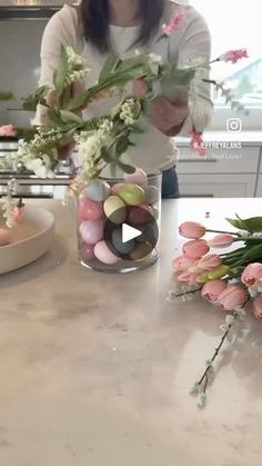 a woman arranging flowers in a vase on top of a kitchen counter next to a plate