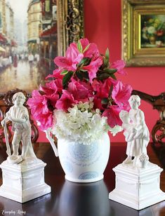 pink and white flowers are in a vase on a table next to two small statues