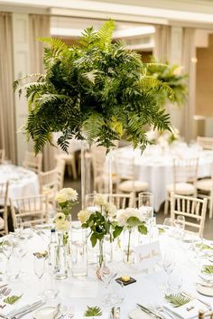 the table is set with white linens, silverware and greenery in tall vases