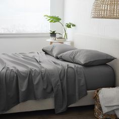 a bed with grey sheets and pillows next to a basket on the floor in front of a window