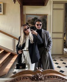 a man and woman taking a selfie in front of a staircase with an ornate handbag