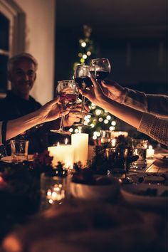 three people toasting wine glasses over a dinner table with candles and christmas tree in the background