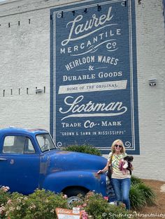 a woman standing next to an old blue truck in front of a sign that says laurel mercantile