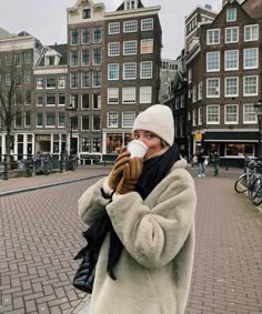 a woman drinking from a cup while standing on a brick walkway in front of buildings