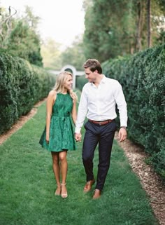 a man and woman holding hands walking down a path in the middle of grass with bushes behind them