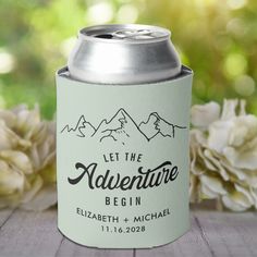 a can of beer sitting on top of a wooden table next to white flowers and greenery