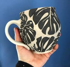 a hand holding a black and white coffee cup with monster leaves on the outside, in front of a blue wall