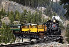 a train traveling over a bridge on top of a forest filled mountain side under a cloudy sky