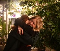 two women hugging each other in front of some trees and greenery at night time