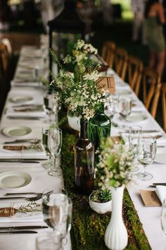 a long table is set with white plates and vases filled with flowers on moss