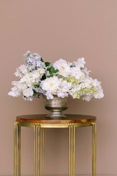 white flowers are in a vase on a gold table with a pink wall behind it