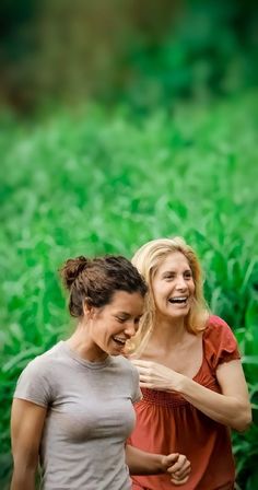 two women laughing and standing next to each other in front of a field of green grass