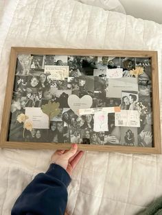 a person holding up a framed photo with pictures and papers attached to it on top of a bed