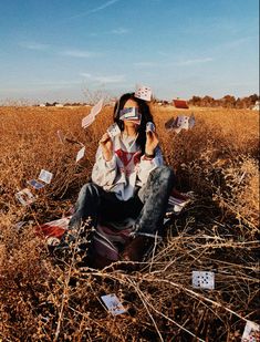 a woman sitting in the middle of a field talking on a cell phone and holding up cards