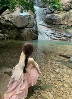 a woman in a dress sitting on rocks near a waterfall and looking at the water