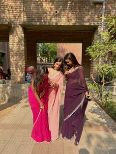 three women in sari posing for the camera on a brick walkway with trees and bushes behind them