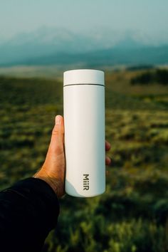 a hand holding a white coffee cup in the middle of a field with mountains in the background