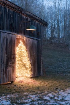 a small christmas tree is lit up in front of a barn with snow on the ground