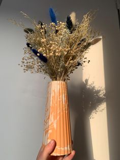 a hand holding a vase with dried flowers in it on a table next to a wall