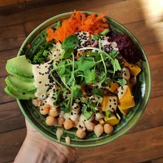 a person holding a green bowl filled with vegetables and fruit on top of a wooden table