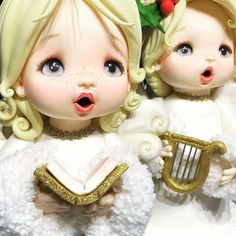 two dolls with blonde hair and blue eyes are dressed in white clothing, holding a golden harp