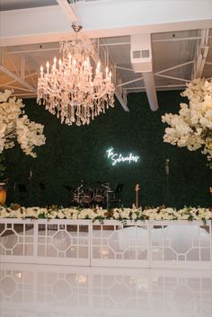 a wedding reception setup with chandelier and flowers on the wall, in front of a green backdrop