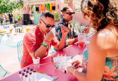 two men and a woman sitting at a pink table with drinks in front of them