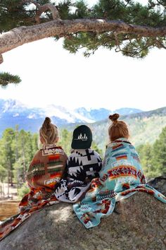 two women sitting on top of a rock under a tree looking out at the mountains