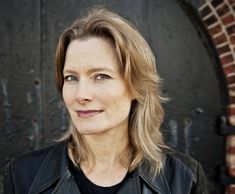 a woman standing in front of a brick wall with a black jacket on and looking at the camera