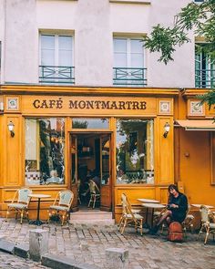 a woman sitting on a bench in front of a yellow cafe with tables and chairs