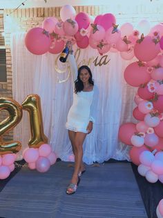 a woman standing in front of a backdrop with pink and gold balloons, holding a champagne bottle