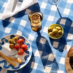 a blue and white checkered table cloth with food on it, including bread, tomatoes, olives, and lemons