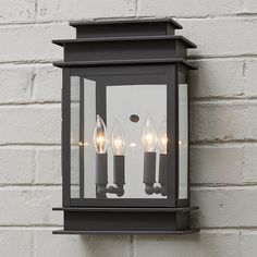 a wall mounted light with three candles on the front and back of it, against a white brick wall