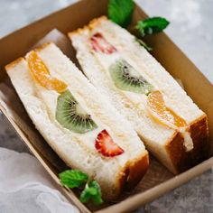 two sandwiches in a box with fruit on top and mint leaves around the edges, being held by someone's hands