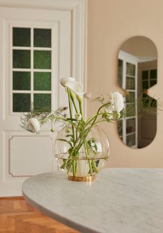 white flowers in a vase sitting on a marble table with a mirror behind it and a wooden floor