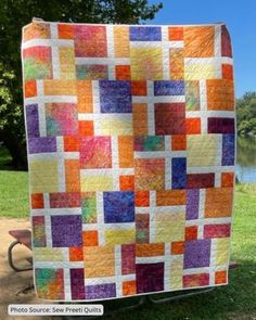 an orange and purple quilt is hanging on a pole in front of some grass with trees