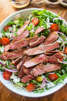 a salad with meat, tomatoes and onions in it on a wooden table next to a fork