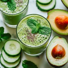 two glasses filled with green smoothie surrounded by sliced cucumbers and mint leaves