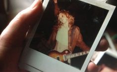 a person holding up a polaroid with an image of a woman playing guitar in the background