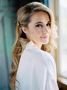 a woman with long blonde hair wearing a white shirt looking out a window at the camera